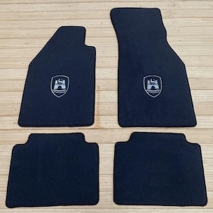 Puede incluir: Un juego de cuatro alfombrillas negras para coche con borde oscuro. Las dos alfombrillas delanteras más grandes presentan un emblema central en forma de escudo con un gráfico de un castillo y líneas onduladas. Las dos traseras son rectángulos negros.