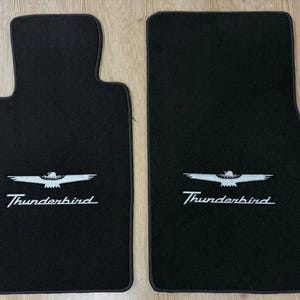 Può includere: Due tappetini per auto neri con il logo e la scritta Thunderbird ricamati in bianco. Il tappetino sinistro ha una forma sagomata, mentre quello destro è rettangolare. Entrambi i tappetini presentano una texture scura e morbida.