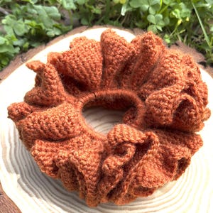May include: A rust-colored, crocheted hair scrunchie featuring a dense, ruffled texture and a circular elastic center. The accessory is crafted with a consistent yarn stitch pattern, creating a voluminous, gathered appearance throughout the entire piece.