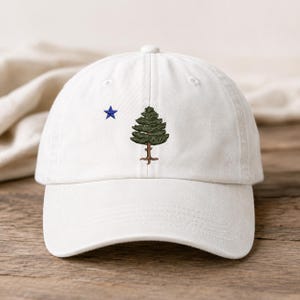 May include: A white baseball cap with a blue star and a green tree embroidered on the front. The cap has a curved brim and a button on top. The hat is made of a soft material and is suitable for casual wear.