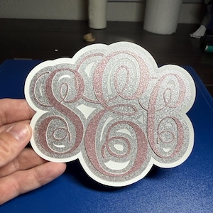 May include: A decorative cutout featuring a three-letter monogram in a swirling script font. The letters are rendered in a rose gold glitter finish, layered over a silver glitter background, all enclosed within a white scalloped border.
