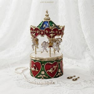 May include: A carousel music box featuring a gold-toned frame with red and green enamel accents. The top canopy is blue and green, while the base displays heart-shaped panels with swan and floral motifs. White horses are suspended from gold poles.