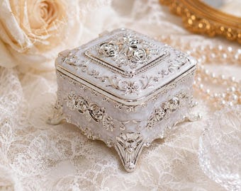 May include: A square white jewelry box featuring intricate silver-toned metal filigree and embossed rose motifs on the lid and sides. The box stands on four ornate, curved silver-toned feet and has a glossy finish with detailed floral patterns.