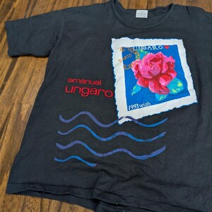 May include: A black short-sleeve t-shirt featuring a graphic of a postage stamp with a red rose and the text "UNGARO", "PARIS", and "1993 wish". The shirt also displays the red text "emanuel ungaro" and blue wavy lines below the stamp graphic.
