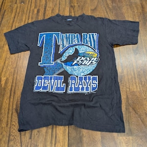 Könnte beinhalten: Ein schwarzes Kurzarm-T-Shirt mit einem großen Grafikdruck. Das Design enthält den Schriftzug Tampa Bay Devil Rays in Blau und Weiß, begleitet von einem kreisförmigen, blau-weiß gemusterten Emblem mit einer schwarzen Rochen-Abbildung.