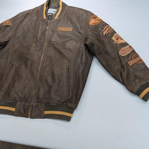 May include: A brown leather bomber jacket featuring a front zipper closure, ribbed collar, cuffs, and hem with yellow stripes. The chest displays the text "LEAK" and the left sleeve is decorated with multiple embroidered patches.