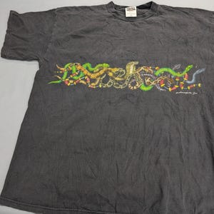 May include: A charcoal grey short-sleeve t-shirt featuring a horizontal graphic across the chest of multiple intertwined snakes in shades of green, yellow, brown, and red. The text Indianapolis Zoo is printed in white near the bottom right of the graphic.