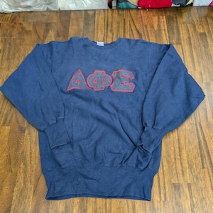 May include: A navy blue long-sleeve sweatshirt featuring the Greek letters Alpha Phi Epsilon in a dark, textured fabric with a red stitched outline across the chest. The garment has a crew neckline, ribbed cuffs, and a ribbed hem at the bottom.
