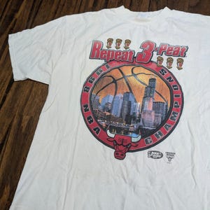 May include: A white short-sleeved t-shirt featuring a graphic print that reads Repeat 3-Peat 1998 NBA Champions. The design includes a basketball, a city skyline, six championship trophies, and a red Chicago Bulls logo at the bottom center.