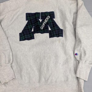 May include: A light gray crewneck sweatshirt featuring a large block letter M applique in a dark green and navy blue plaid pattern. The word GOPHERS is embroidered in white across the letter M. A small Champion logo is on the left sleeve cuff.
