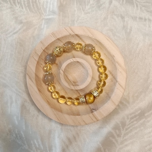 May include: A bracelet with a mix of golden and grey gemstones. The bracelet includes decorative gold-coloured beads and is displayed on a wooden coaster. The bracelet is a stylish accessory.