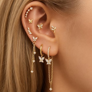 May include: A collection of gold-toned earrings featuring clear cubic zirconia stones. The set includes a curved climber, a small butterfly stud, a leaf-shaped stud, and two hoop earrings with dangling butterfly charms and delicate chain accents.