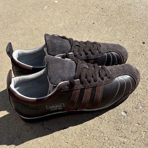 May include: A pair of dark brown Adidas Cup 68 sneakers featuring a suede toe cap and tongue, leather side panels with three brown suede stripes, white stitching details, a light gray interior lining, and a black sole with a white midsole stripe.