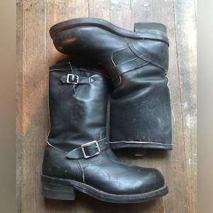 May include: A pair of black leather engineer boots featuring a rounded toe, a low stacked heel, and two adjustable straps with silver-toned metal buckles. One strap is positioned across the instep, while the other is located at the top shaft.