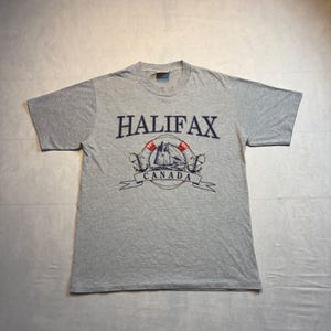以下が含まれることがあります： ヘザーグレーの半袖Tシャツ。濃い青色の文字で「HALIFAX」と書かれ、その下に赤いアクセントの入った救命浮環の中の帆船のグラフィックと2つの錨が描かれ、さらにその下のバナーに「CANADA」という文字が記されています。