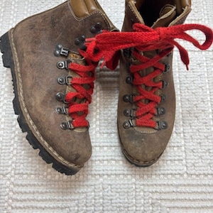 May include: A pair of brown leather hiking boots featuring a high-top design with padded collars, dark metal D-ring eyelets, thick black rubber lug soles, and bright red textured laces that are tied in a bow at the top of the boots.
