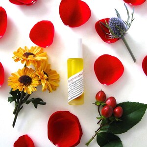 May include: A small glass bottle of yellow facial oil with a white label that reads "Regenerative Anti-Aging Intensive Serum". The bottle is surrounded by red rose petals, yellow flowers, and green leaves.