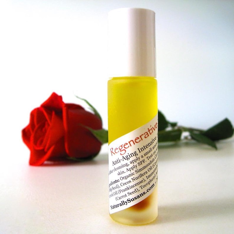 May include: A clear glass bottle with a yellow liquid inside. The bottle has a white cap and a label that reads "Regenerative Anti-Aging Intensive Serum". The label also lists ingredients, including organic Simmondsia Chinensis Seed Oil (Jojoba Seed), Cocos Nucifera Oil (Coconut Oil), Frankincense, Immortelle (Carrot Seed), and Tocopheryl (Vitamin E). The bottle is sitting on a white surface next to a red rose.