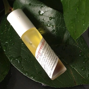 May include: A white and brown glass roll-on bottle with a label that reads "Regenerative Anti-Aging Intensive". The bottle is on a green leaf with water droplets.