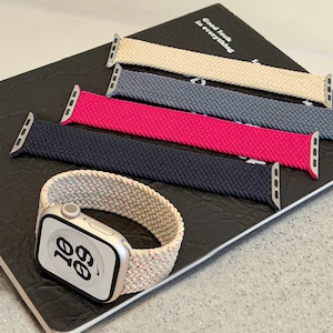 Puede incluir: Una colección de cuatro correas de reloj trenzadas en beige, gris, magenta y azul oscuro, junto a un reloj inteligente plateado con pantalla blanca que muestra la hora 10:09 y una correa trenzada beige con sutiles motas rojas unida al dispositivo.