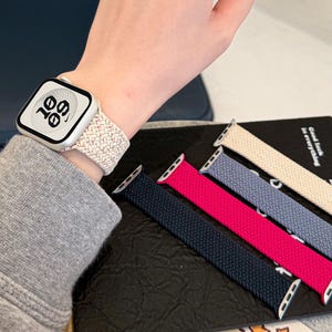Peut inclure: Une montre connectée argentée avec un écran numérique affichant 10:09 et un bracelet élastique tressé blanc avec de fines taches rouges. Quatre bracelets tressés supplémentaires en beige, bleu, magenta et bleu marine sont présentés à côté.