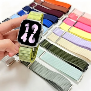 Puede incluir: Un reloj inteligente rectangular con pantalla negra que muestra la hora 14:46 en grandes números blancos, sujeto a una correa de nailon acanalada de color verde lima. Varias correas de reloj adicionales de diversos colores están dispuestas al fondo.