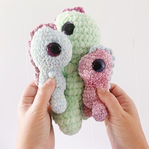 Può includere: Tre creature all'uncinetto fatte a mano nei toni del blu, verde e rosa. Ognuna ha un grande occhio scuro e un corpo strutturato. Le creature sono tenute su uno sfondo bianco.