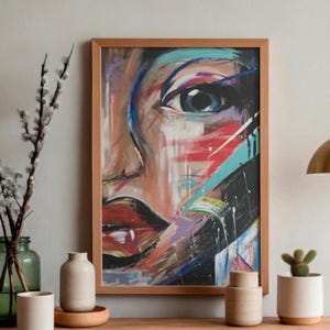 May include: A rectangular framed wall art print featuring an abstract, expressive painting of a human face. The artwork uses bold, gestural brushstrokes in shades of red, black, teal, and white, focusing on a prominent eye and dark, defined lips.