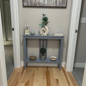 May include: A blue console table with two levels, featuring a white lantern, a gray sphere, a cream-colored swirl vase with teal flowers, and a small book on top. The lower shelf holds a woven bowl, a small potted topiary, and a white bowl.
