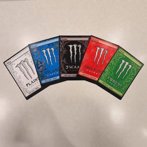 May include: Five custom trading cards with the Monster Energy logo. The cards are labeled Plains, Island, Swamp, Mountain, and Forest. Each card features a unique color scheme and design, with the Monster Energy logo prominently displayed.