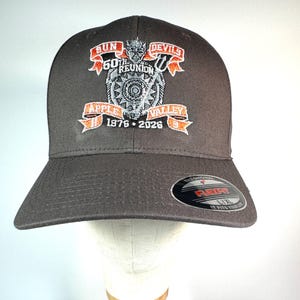 May include: A dark gray baseball cap featuring an embroidered graphic with the text SUN DEVILS 60TH REUNION APPLE VALLEY 1976-2026 and a trident-wielding mascot. A black and silver sticker on the brim reads THE ORIGINAL FLEXFIT L/XL IT FITS YOUR LIFE.