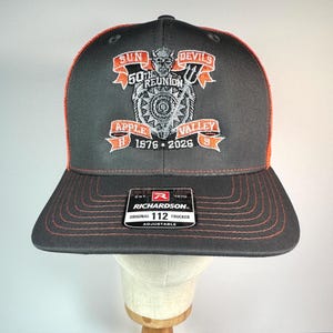 May include: A charcoal gray trucker hat with orange stitching and an embroidered patch featuring a devil mascot, a shield, and text reading Sun Devils 50th Reunion Apple Valley H S 1976-2026. A black Richardson 112 Original Adjustable sticker is on the brim.