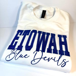 May include: A white crewneck sweatshirt featuring the text ETOWAH in large blue block letters and Blue Devils in blue cursive script embroidered across the chest. The garment has a black neck label and is folded on a plain white surface.
