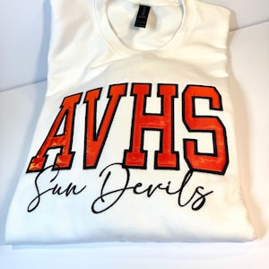 May include: A white crew neck t-shirt featuring the text AVHS in large orange block letters with black outlines. Below the text, the words Sun Devils are printed in a black cursive script font. The shirt has a standard collar and short sleeves.