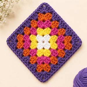 May include: A square crochet granny square featuring a central white flower motif surrounded by concentric layers of yellow, pink, orange, and purple yarn. The pattern consists of repeating clusters of stitches forming a geometric floral design.