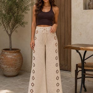 May include: A dark brown ribbed tank top paired with beige crochet wide-leg pants. The pants feature a drawstring waist and vertical side panels with a repeating cutout flower pattern that runs down the length of each leg to the hem.