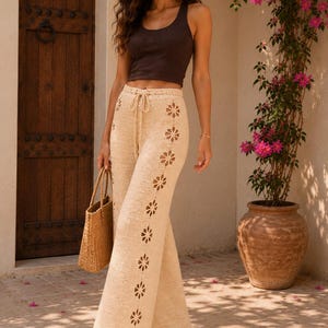 May include: A dark brown ribbed tank top paired with cream-colored crochet wide-leg pants featuring a vertical row of floral cutouts along the side and a drawstring waist. The outfit is completed with a woven straw tote bag and tan sandals.