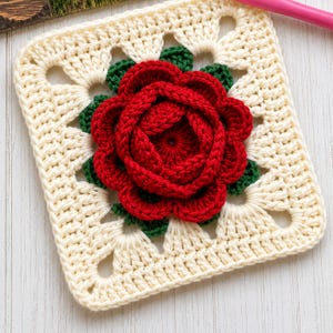 May include: A square, cream-colored crochet piece with a red rose in the center. The rose is surrounded by green leaves. The square has a decorative openwork border, showcasing a handmade craft.