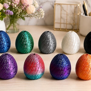 May include: A collection of ten decorative egg-shaped objects in various colors, including blue, green, gray, white, black, purple, red, and orange. Each egg has a textured surface with a vertical pattern. The eggs are arranged on a light-colored surface.