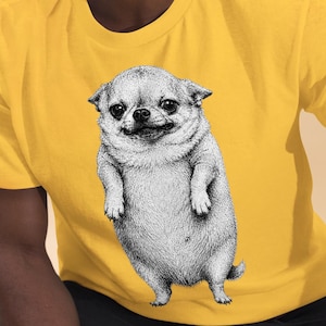 Puede incluir: Una camiseta de manga corta de color amarillo brillante con una ilustración detallada en blanco y negro, estilo tinta, de un perro pequeño y robusto de pie sobre sus patas traseras, centrada en el pecho. El gráfico muestra un perro de cuerpo redondeado.