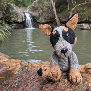 May include: A handmade crochet plush toy shaped like a dog with grey, black, and tan yarn. The dog features pointed ears, black plastic eyes, a black nose, and stitched paw details on its four feet. The toy is crafted with a dense, textured stitch.