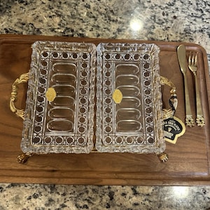 May include: Two rectangular crystal serving dishes with gold accents and handles sit on a wooden tray. A gold knife and fork are to the right. A tag reads "24K GOLD PLATED".