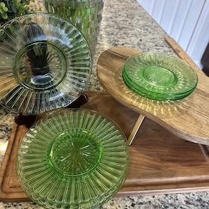 May include: A collection of green glass plates with a ribbed design, displayed on wooden serving boards. The plates are arranged in a visually appealing manner, showcasing their intricate details and vibrant color. The plates are a vintage style.