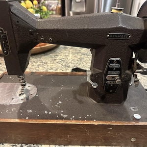 May include: Vintage Kenmore sewing machine in dark brown with a silver needle and rotary controls. The machine is mounted on a wooden base. The Kenmore logo is visible on the machine.
