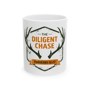 May include: A white ceramic mug featuring a dark green graphic of deer antlers framing a shield shape. The text reads THE DILIGENT CHASE in orange, with PROVERBS 12:27 printed in black on an orange banner at the bottom of the shield design.
