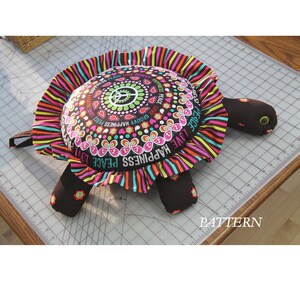 Pode incluir: Uma almofada de tartaruga colorida feita à mão com um sinal de paz e as palavras "Peace, Love, Happiness" na casca. A almofada é feita de tecido com fundo preto e listras coloridas.