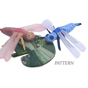 Peut inclure: Deux jouets en tissu en forme de libellule avec des ailes roses et bleues, posés sur un nénuphar en tissu vert. Le texte "PATTERN" est visible sous le nénuphar.