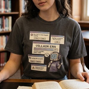 May include: A charcoal gray short-sleeve t-shirt featuring a graphic print of computer error pop-up windows, a loading bar labeled VILLAIN ERA, and a sad robot character with a monitor head wearing headphones and a purple shirt.