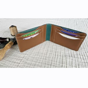 Peut inclure: Portefeuille en cuir marron ouvert, avec des accents turquoise et des coutures blanches. Le portefeuille est ouvert, révélant plusieurs fentes pour cartes et une tranche turquoise. Sur une surface blanche texturée, avec des outils de maroquinerie.
