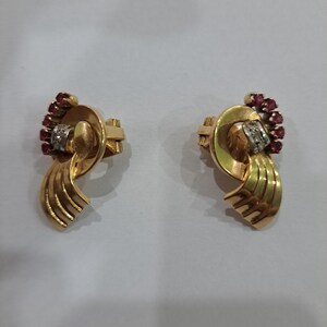 May include: A pair of gold-toned earrings with a swirling design. Each earring features a cluster of small red gemstones and a silver-toned accent. The earrings have a clip-on style.
